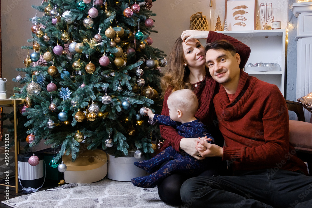 Beautiful young family sits on a carpet near the fireplace next to a large elegant Christmas tree Beautiful young family sits on a carpet near the fireplace next to a large elegant Christmas tree