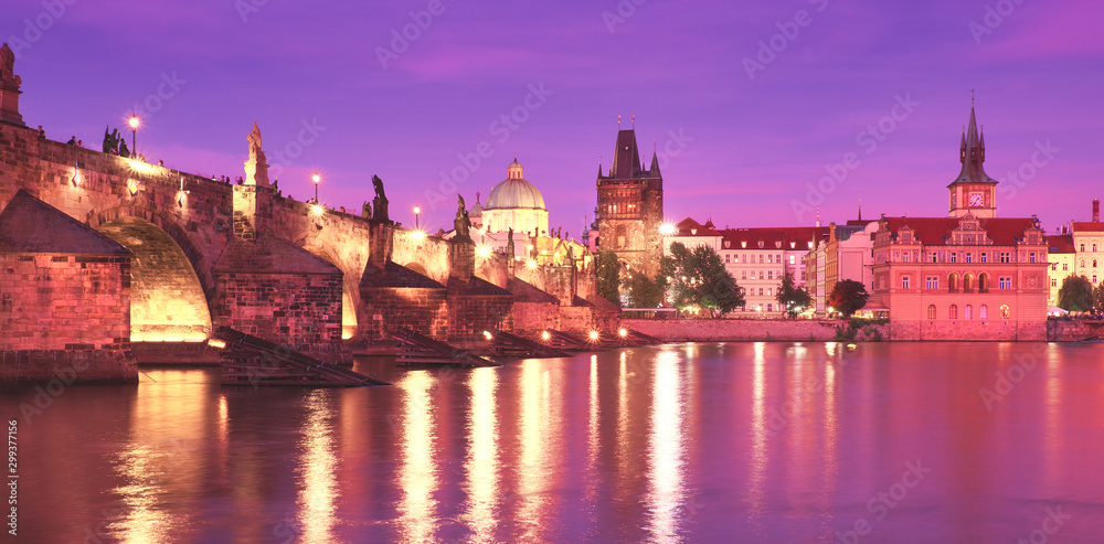 Prague on sunset, illuminated Charles bridge, Bridge Tower and old buildings on the riverside under purple sky with golden reflections in Vltava river, panoramic image