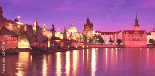 Prague on sunset, illuminated Charles bridge, Bridge Tower and old buildings on the riverside under purple sky with golden reflections in Vltava river, panoramic image