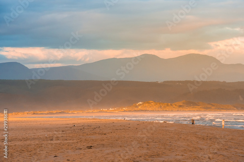 A streak of golden evening light falling on the Plettenberg Bay beach at sunset, with mountains in the distance and people walking on the beach. Garden Route, Western Cape, South Africa