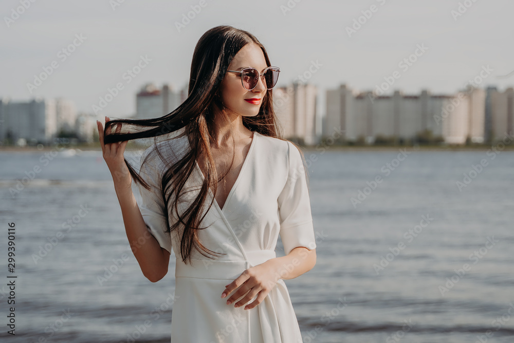 young woman by the river. Urban landscape. Brunette in a white dress. Long hair. Beauty and fashion. Girl in sunglasses young woman by the river. Urban landscape. Brunette in a white dress. Long hair. Beauty and fashion. Girl in sunglasses