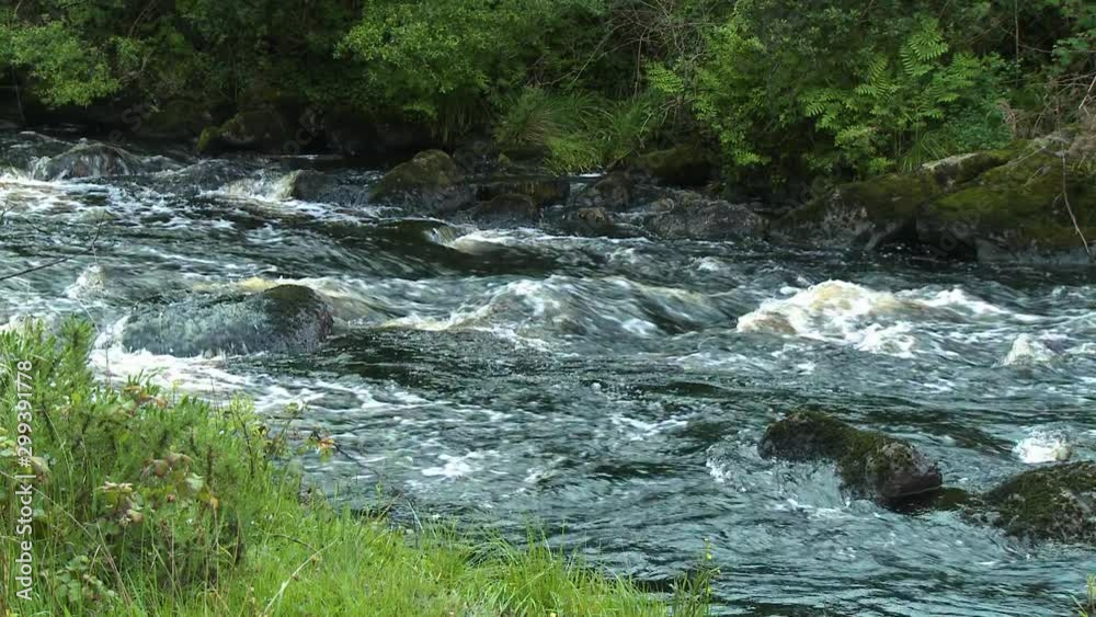 Steady, medium wide shot of a fast flowing small river and a grassy bank.