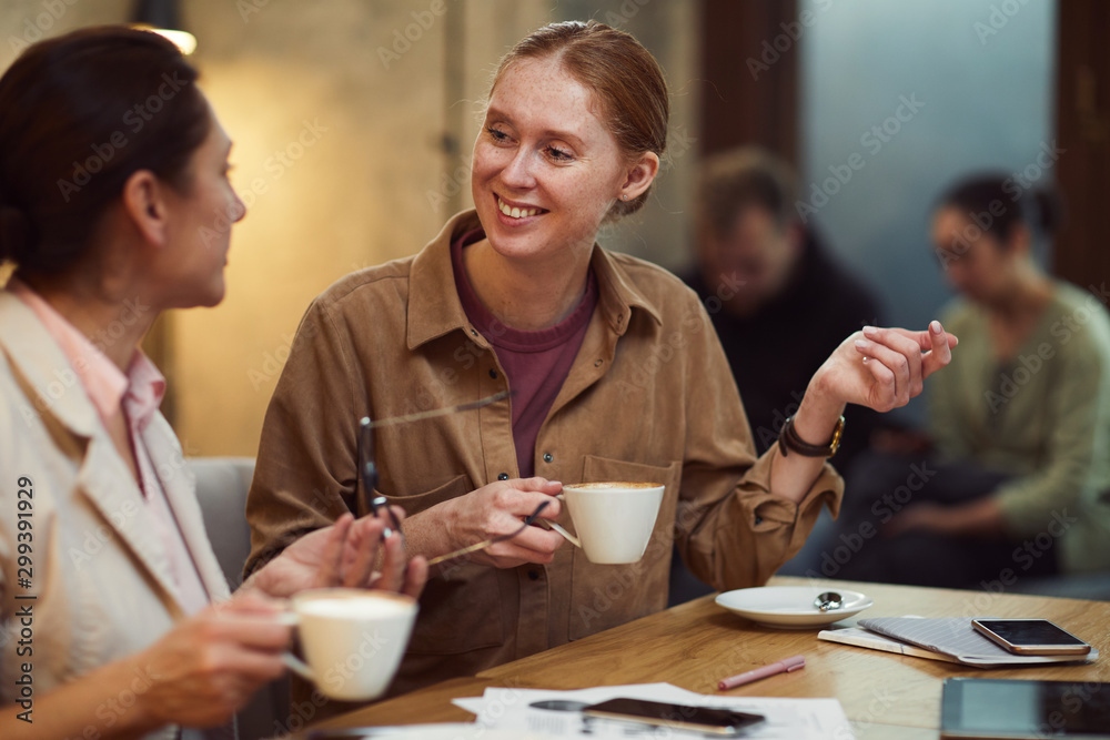 Young smiling businesswoman talking to her colleague at the table while they drinking coffee at cafe