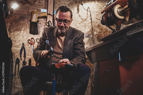 Focused mature cobbler is fixing sole for boots using hammer. © Fxquadro