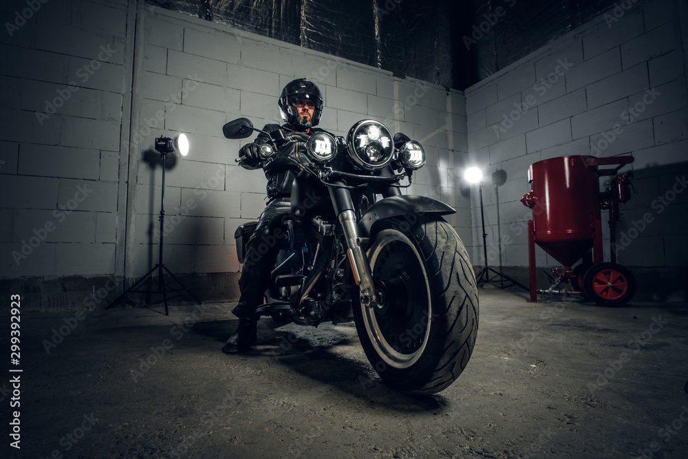 In dark garage bearded biker on motorbike is posing for photographer on his bike.