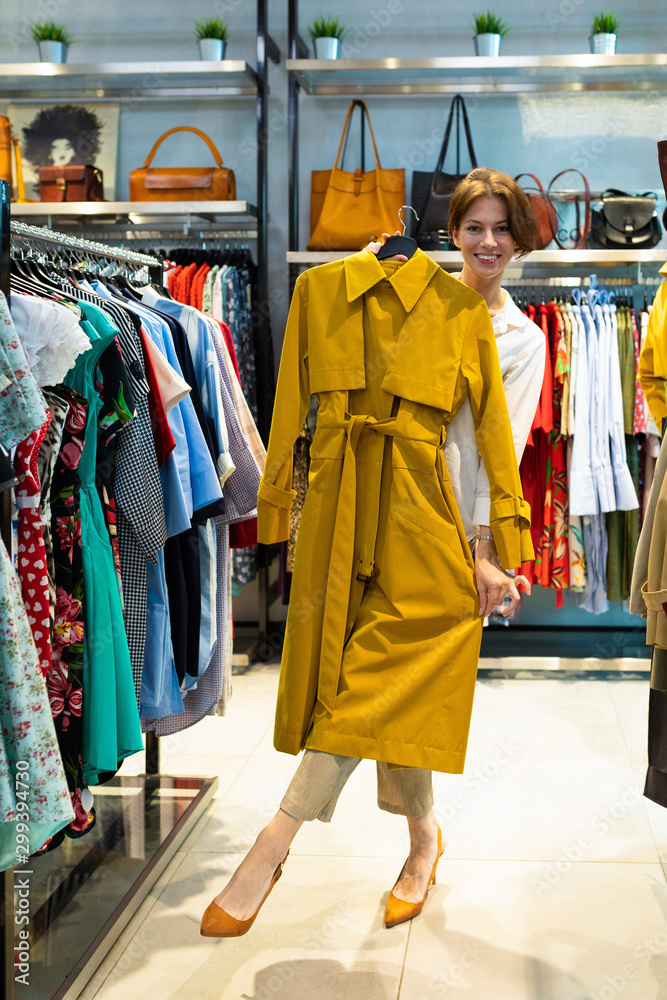 Young woman is smiling and demonstrating raincoat of mustard color in clothing shop for women