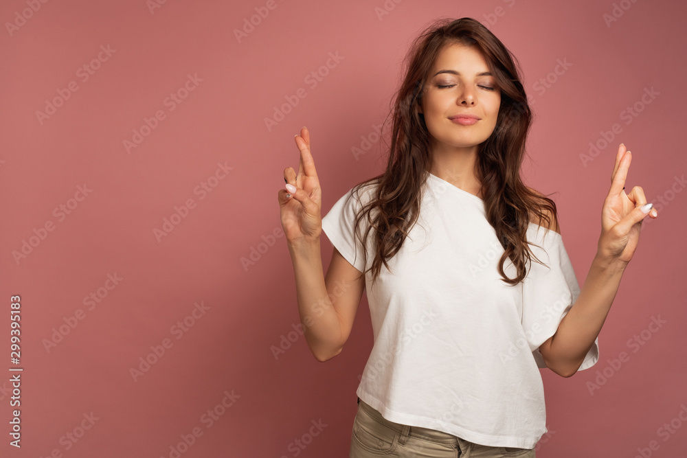 A dark-haired girl in a white top is standing on a pink background with closed eyes and crossed fingers. A dark-haired girl in a white top is standing on a pink background with closed eyes and crossed fingers.