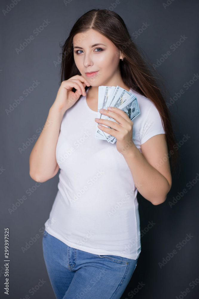 young, beautiful girl in a white T-shirt is holding dollars in hands. Studio photo on a gray background. young, beautiful girl in a white T-shirt is holding dollars in hands. Studio photo on a gray background.