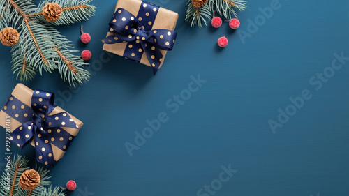 Fototapeta Naklejka Na Ścianę i Meble -  Christmas tree branch with red berries and gift box over blue xmas background. Flat lay, top view, copy space. Christmas banner mockup with copy space
