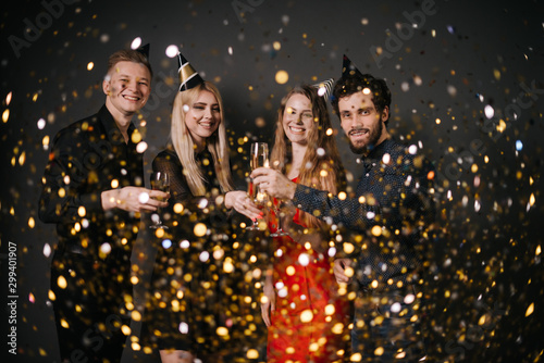 Two attractive guys and two beautiful girls are clinking with champagne glasses and celebrating new year. On friends from above the festive confetti falling. Shooting on isolated black background.