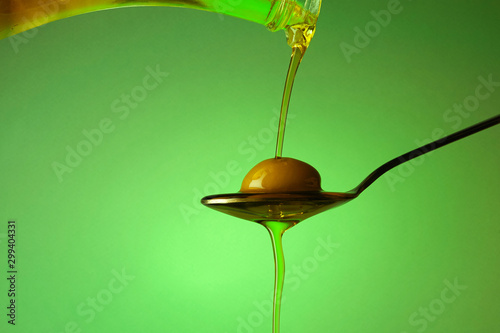 A spoon with an olive under a fall of oil on green background