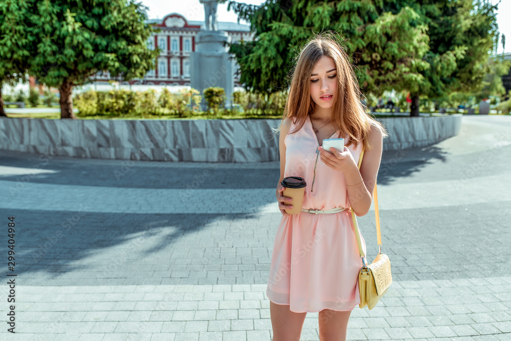 girl in pink dress, stands in city in summer, looks at phone, reads a message online on Internet, a cup of coffee tea, social networking application. Free space for text.