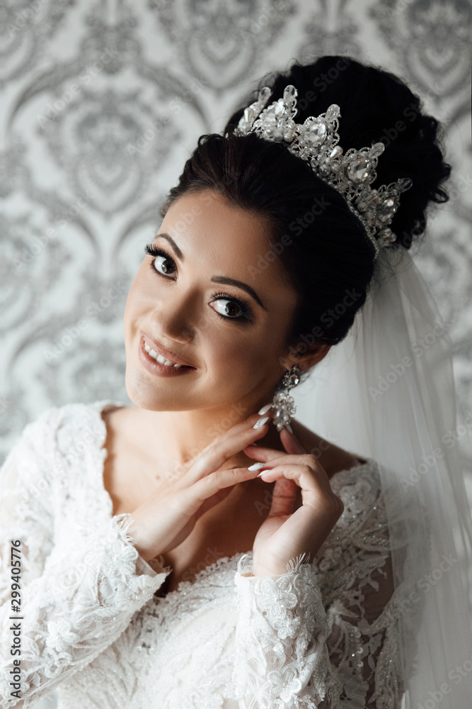 Very beautiful bride. Pretty brunette bride with hair style, sensual make up and diadem on her head. Beautiful charming bride touches earrings. Beautiful and fashion bride in home Very beautiful bride. Pretty brunette bride with hair style, sensual make up and diadem on her head. Beautiful charming bride touches earrings. Beautiful and fashion bride in home