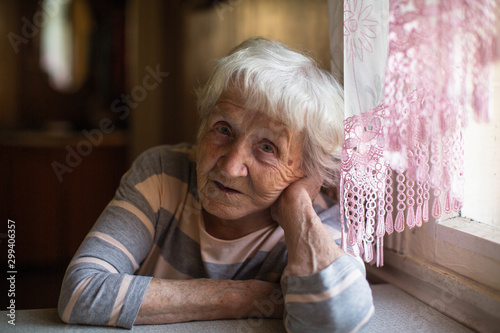 Elderly woman sits sadly near the window at his home.