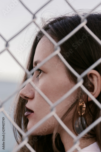 Pretty woman stands against wired fence. Model in white shirt. Purple lips. Pretty make up. Posing for magazine. Street fashion. Urban lifestyle. lady with long hair, beautiful eyes, behind fence.