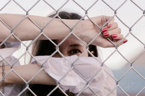 Pretty woman stands against wired fence. Model in white shirt. Purple lips. Pretty make up. Posing for magazine. Street fashion. Urban lifestyle. lady with long hair, beautiful eyes, behind fence.