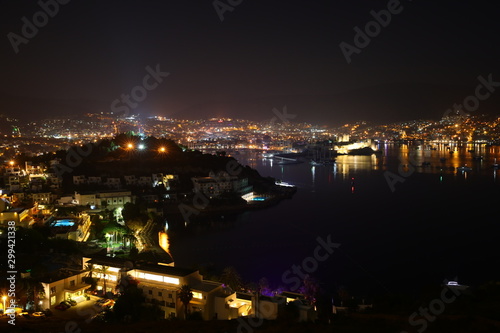 View from the hill to the colorful panorama of the night Bodrum mountains Marina attractions Turkey.Multi-colored neon lights,lamps,lights, night resort.Reflection in water Background Wallpaper © Nina