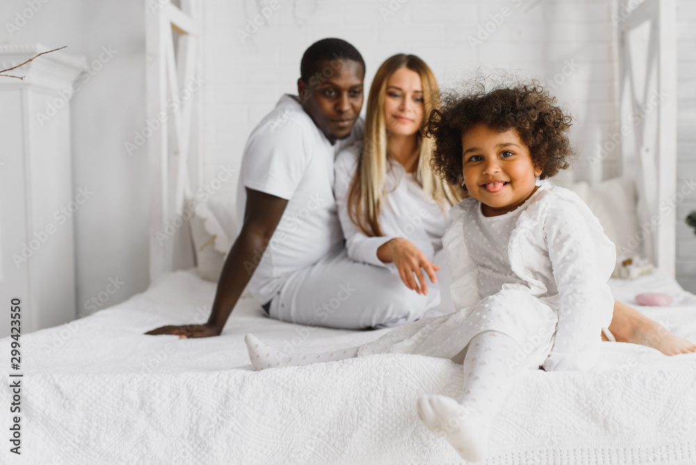 Portrait of happy multiracial young family lying on cozy white bed at home, smiling international mom and dad relaxing with little biracial girl child posing for picture in bedroom Portrait of happy multiracial young family lying on cozy white bed at home, smiling international mom and dad relaxing with little biracial girl child posing for picture in bedroom
