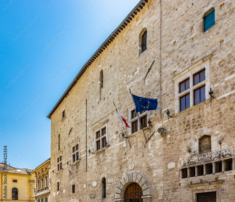 Narni, Umbria, Italy - A large fountain in the center of the village of Narni. The old brick town hall building. Blue sky in the summer. The flag of Italy and the European Community (EEC
