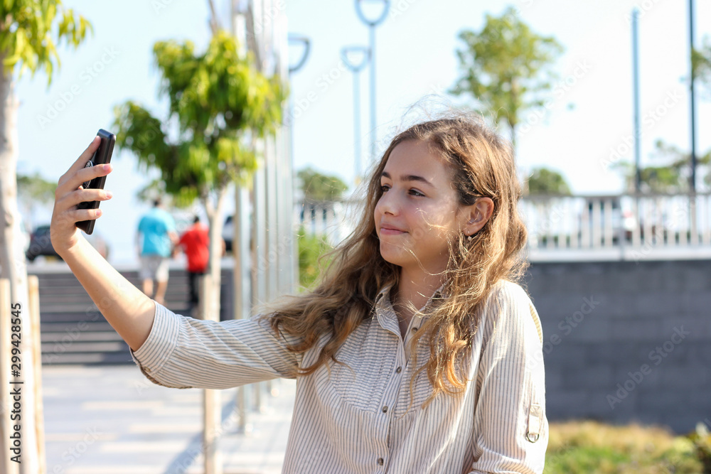 Beautiful teenager girl taking a selfie on the promenade on a sunny day. Happy young girl smiling with a phone in her hands