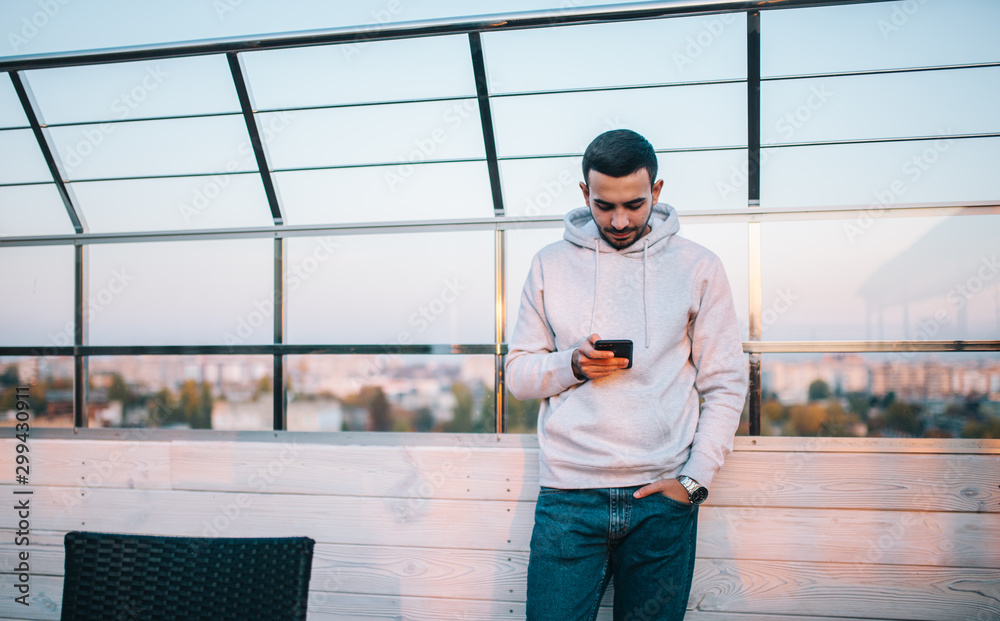 Young man in gray hoodie uses smartphone sits in cafe and reads news, checks e-mail. E-news concept. Young man in gray hoodie uses smartphone sits in cafe and reads news, checks e-mail. E-news concept.