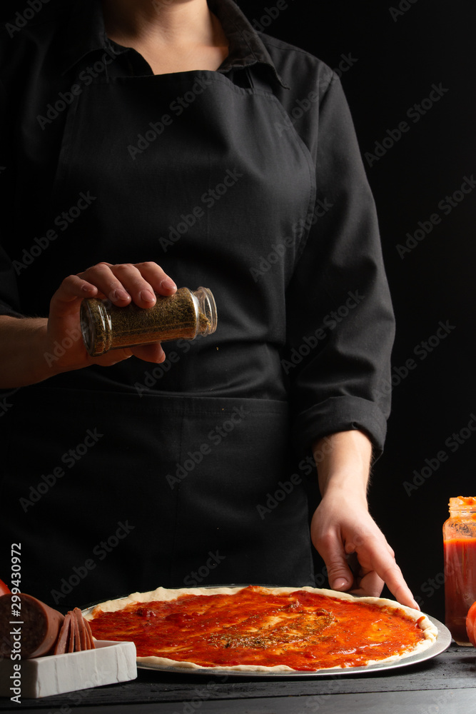 Chef cooks Italian pizza, sprinkled with oregano. Freeze in the air. Against the background of pizza ingredients. Black background, vertical frame.