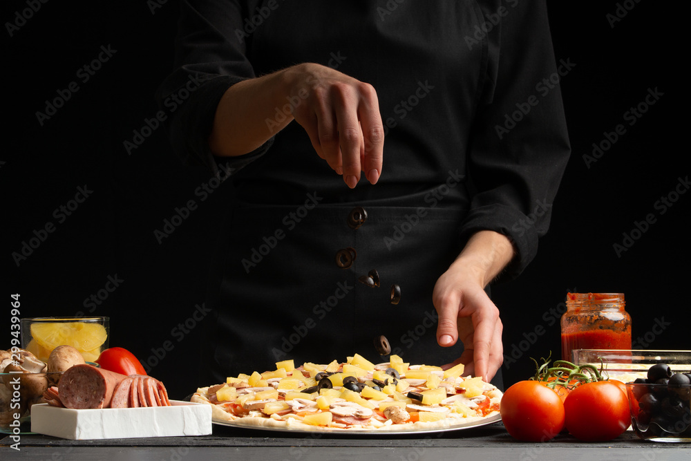 Chef cooks Italian pizza, sprinkles olives. Freezing in motion. Against the background of pizza ingredients. Black background, pizzeria, recipe book, menu, restaurant business.