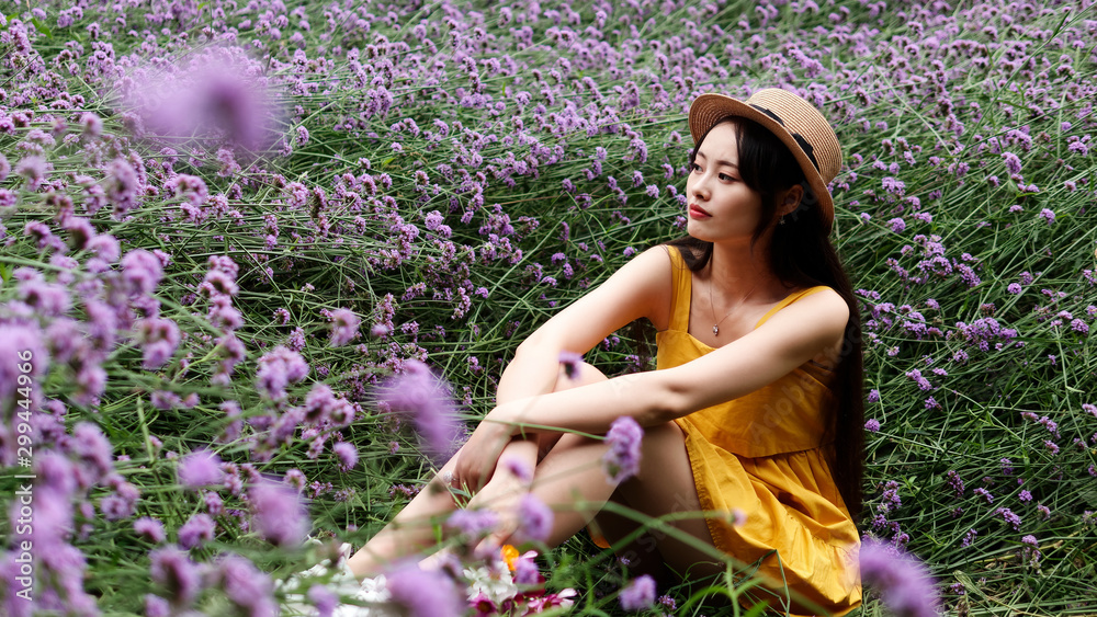 Beautiful woman in yellow dress and straw hat sitting in purple Verbena Bonariensis flower field, charming Chinese girl with black long hair enjoy her leisure time outdoor.