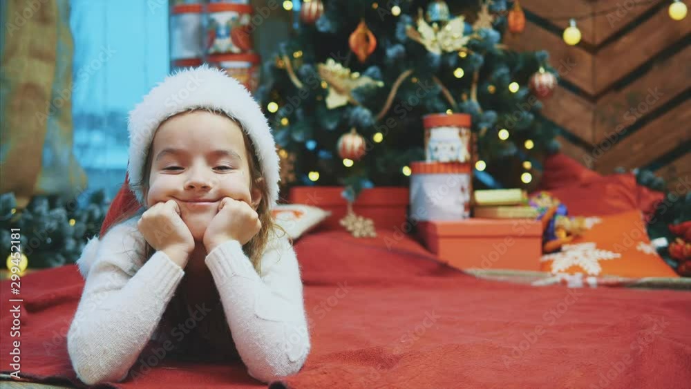 Funny video where little kid lies on red blanket under fir-tree, making comic face expression, looking like cute gnome, weared in red santa hat. Funny video where little kid lies on red blanket under fir-tree, making comic face expression, looking like cute gnome, weared in red santa hat.