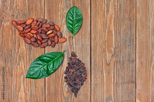 Green leaves and two leaves made of cocoa beans and cacao nibs on wooden background. Top view. Text space. Ecology concept.