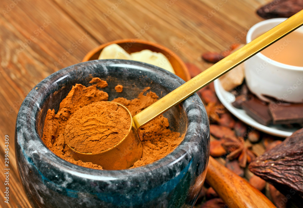 Fototapeta premium Cocoa powder in stone mortar with cup of cacao drink, cocoa pod, chocolate bar, cocoa beans and star anise on wooden background.