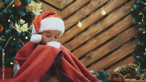 Close-up video of sleepy child in santa hat, wrapped in red blanket near christmas tree, with a cup of cocoa, looking delighted and pleased. photo