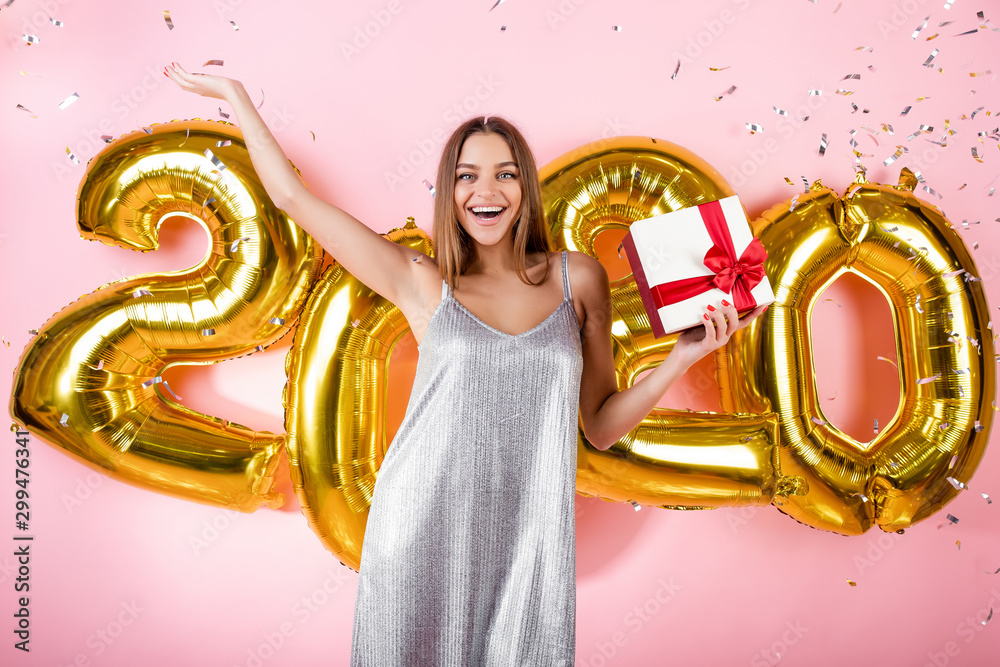 happy woman with gift box and silver confetti and golden 2020 new year balloons isolated over pink