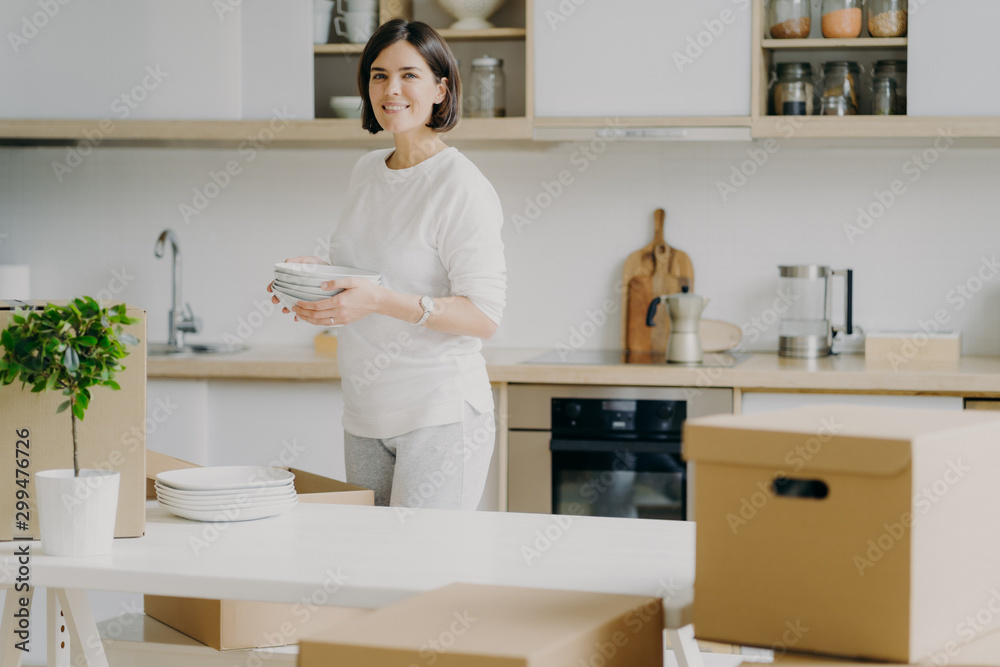 Happy brunette woman in casual outfit, moves into new house, unpackes boxes with kitchen dishes, holds plates, stands near table, green indoor plan in pot, enjoys domestic atmosphere. Modern apartment Happy brunette woman in casual outfit, moves into new house, unpackes boxes with kitchen dishes, holds plates, stands near table, green indoor plan in pot, enjoys domestic atmosphere. Modern apartment