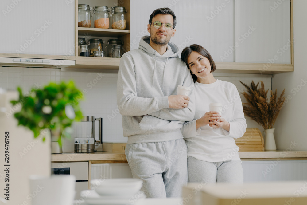 Shot of relaxed family couple stand in kitchen, drink takeaway coffee, wear casual outfit, pose in kitchen interior, bought new apartment for living. People, real estate and mortgage concept Shot of relaxed family couple stand in kitchen, drink takeaway coffee, wear casual outfit, pose in kitchen interior, bought new apartment for living. People, real estate and mortgage concept