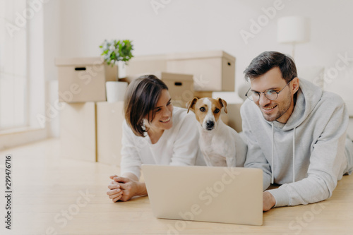 People and new apartment concept. Happy young husband and wife relax on floor, use modern laptop computer for surfing net, pose in their new home or flat, their dog poses near, have little rest