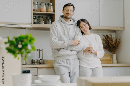 Shot of relaxed family couple stand in kitchen, drink takeaway coffee, wear casual outfit, pose in kitchen interior, bought new apartment for living. People, real estate and mortgage concept
