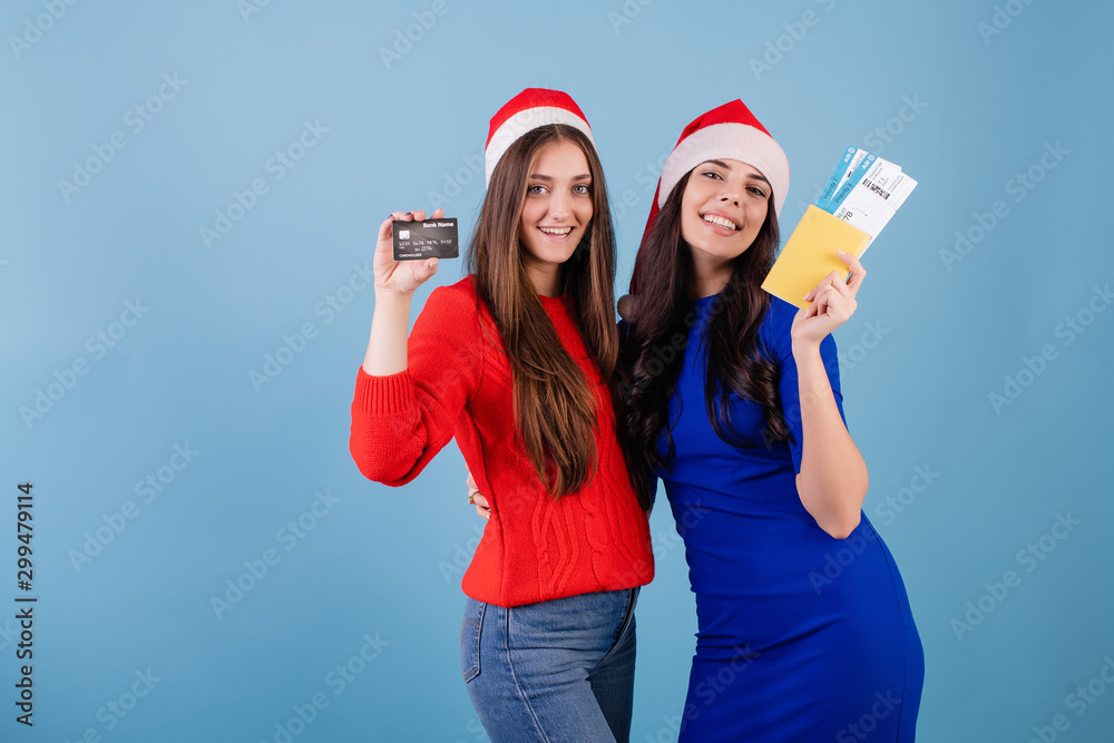 two women wearing santa hats with airplane tickets and passport and credit card isolated over blue two women wearing santa hats with airplane tickets and passport and credit card isolated over blue