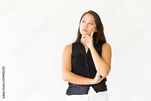 Pensive serious female customer touching face with finger and looking at copy space. Young Latin woman in casual standing isolated over white background. Advertising concept