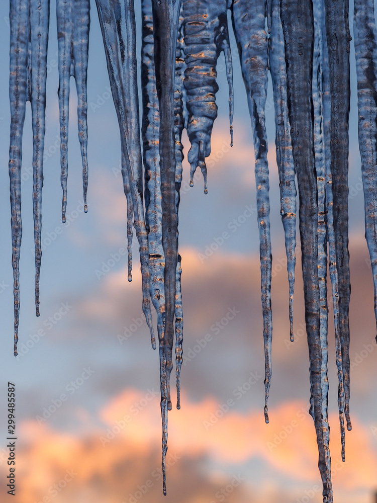 Ice icicles hanging from roof of house. Window covered with large and dangerous, but beautiful transparent icicles. Blue sky with clouds background.