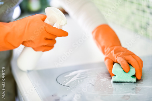 Woman cleaning modern white electric stove with orange rubber gloves. Washing induction cooker using spray bottle And sponge.