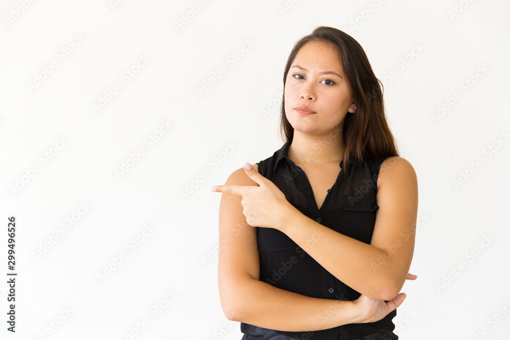 Serious young woman pointing away. Beautiful confident young woman pointing aside with finger and looking at camera. Gesture concept Serious young woman pointing away. Beautiful confident young woman pointing aside with finger and looking at camera. Gesture concept