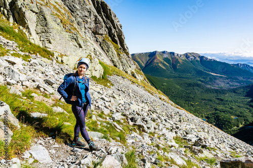 Young attractive tourist with a backpack and a helmet on a slope in the mountains on a beautiful sunny day. In the background a mountain ridge and a valley are visible.