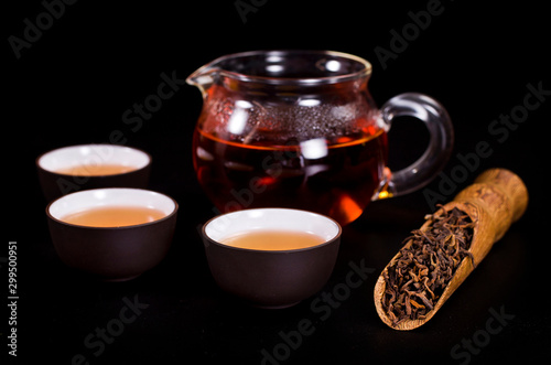 Ceramic tea cups, glass jug of tea with dried black tea in a scoop on a black background.