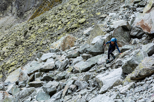 A young mountaineer (woman) in a helmet, with a backpack and trekking poles descends carefully along the path between the large boulders in the valley in the mountains.