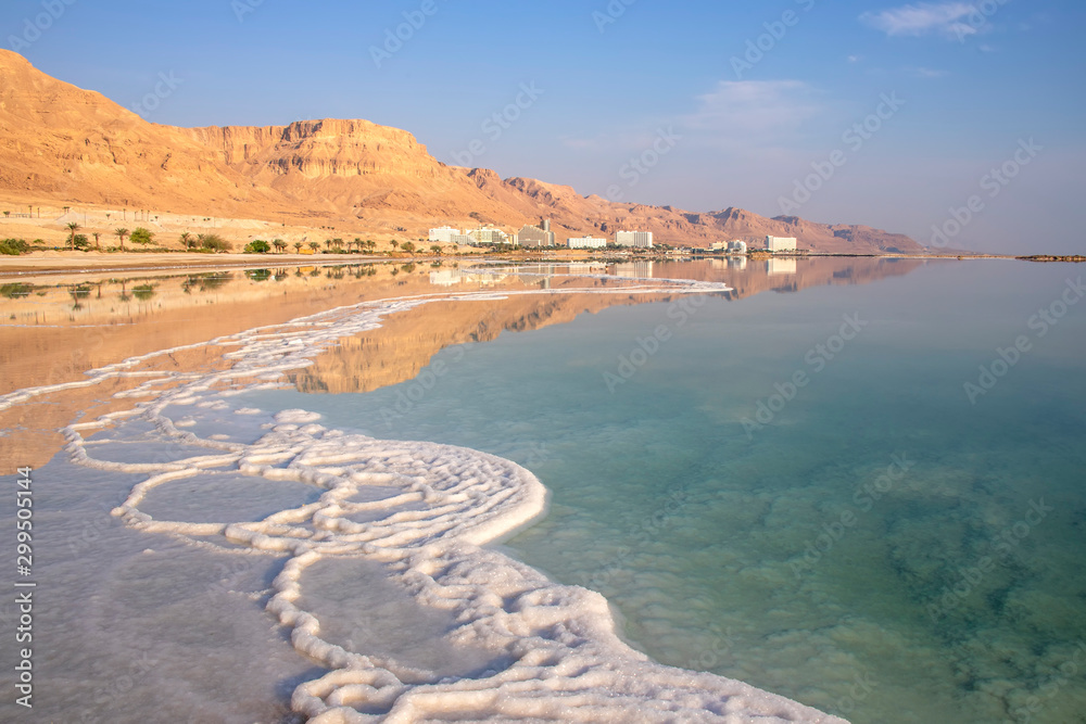 Reflection of mountains, hotels and palm trees in the water of the Dead Sea with salt formations