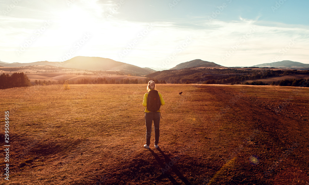 Female hiker with backpack standing on meadow looking at mountain enjoying the view during the sunset.