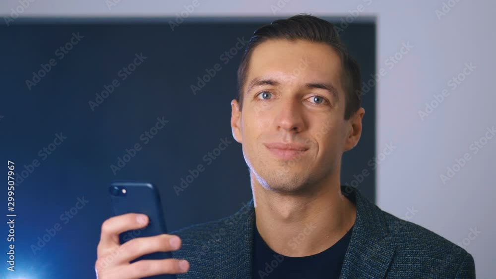 Young Handsome Businessman using his Smartphone inside office background, Looking at Camera and Smiling. Business Lifestyle.