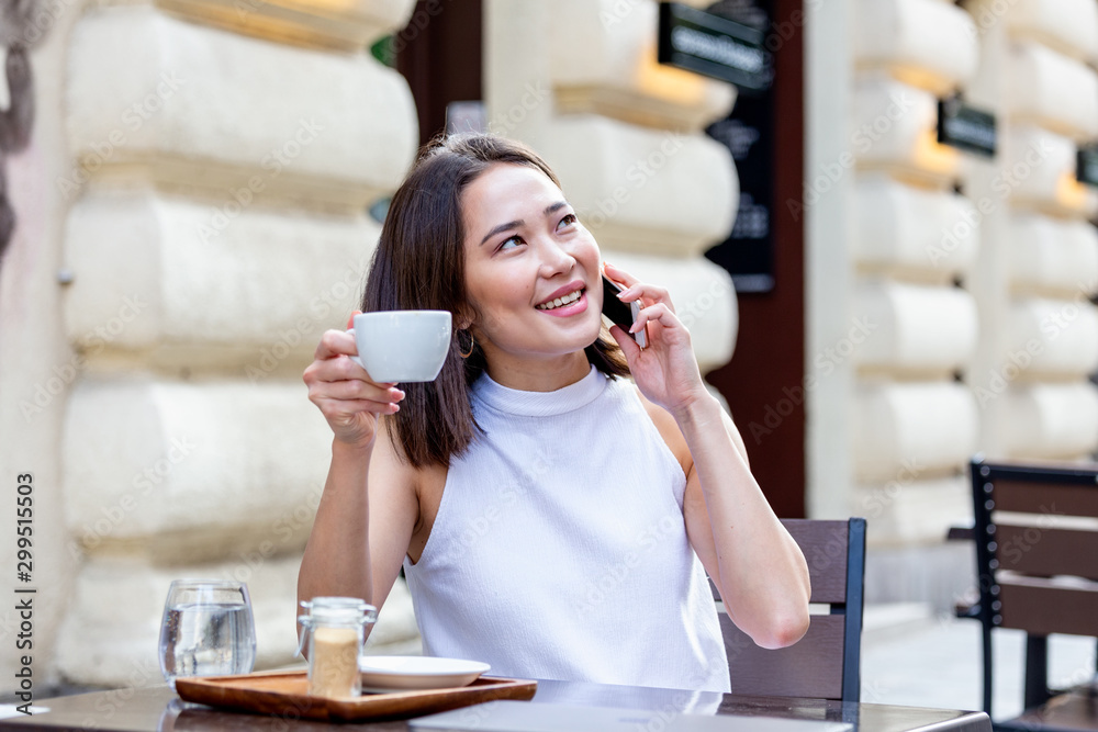 Young Asian woman sitting in coffee shop at wooden table, drinking coffee and using smartphone.On table is laptop. Girl browsing internet, chatting, blogging. Young Asian woman sitting in coffee shop at wooden table, drinking coffee and using smartphone.On table is laptop. Girl browsing internet, chatting, blogging.