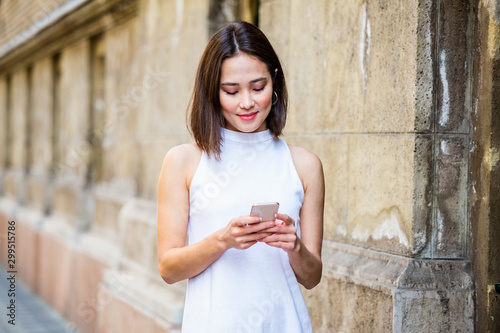 Beautiful asian woman using an application to send an sms message in her smartphone device. Happy young Asian woman with smartphone standing in the street
